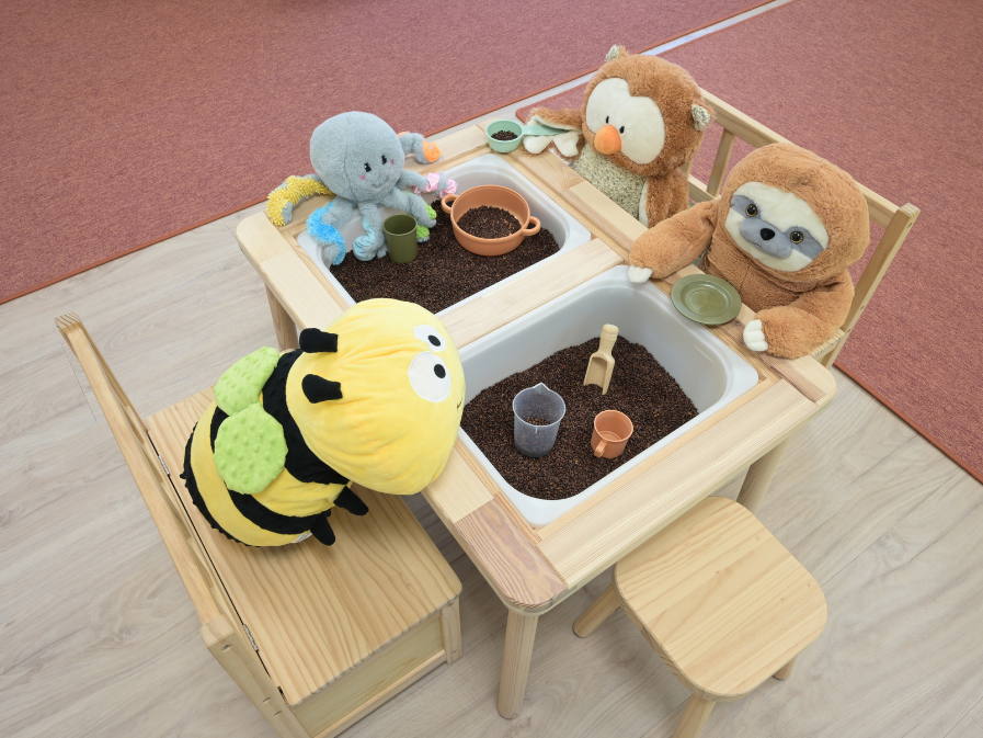 Fühlwanne mit Traubenkernen Spielbereich mit Kuscheltieren: Tintenfisch, Biene, Faultier und Eule an Tisch mit Traubenkernen und Spielzeug.